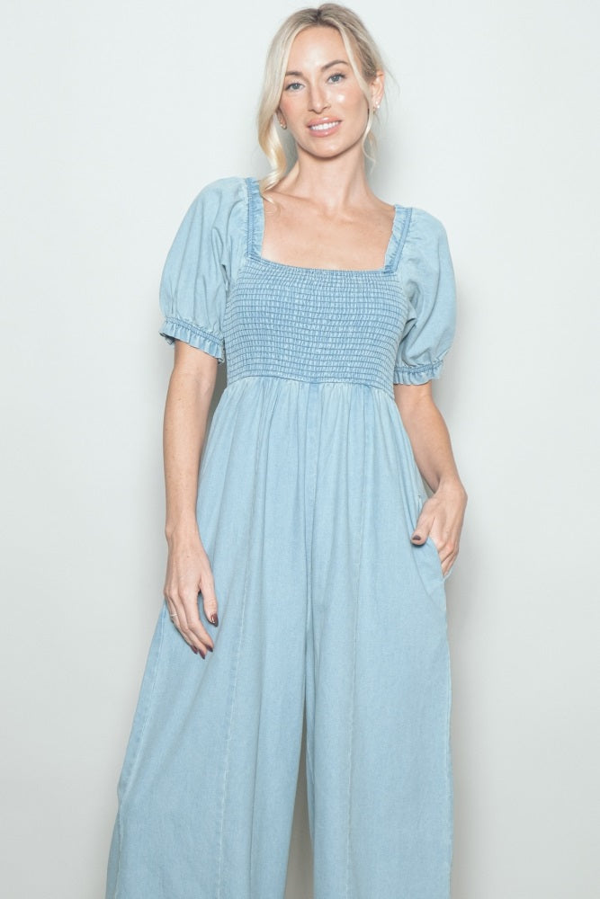 woman wearing a light blue smocked waist jumpsuit with puffy sleeves and side pockets