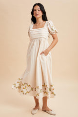 woman wearing a cream-colored midi dress with puff sleeves and floral embroidery on the hem, posing with hands in pockets