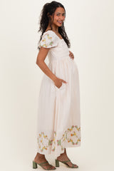 woman wearing a cream maternity maxi dress with floral embroidery and puff sleeves, holding her baby bump