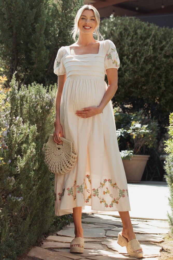 woman wearing a cream maternity dress with short puff sleeves and floral embroidery, holding a woven bag and showing her baby bump