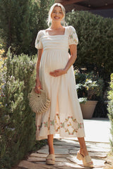 woman wearing a cream maternity dress with short puff sleeves and floral embroidery, holding a woven bag and showing her baby bump