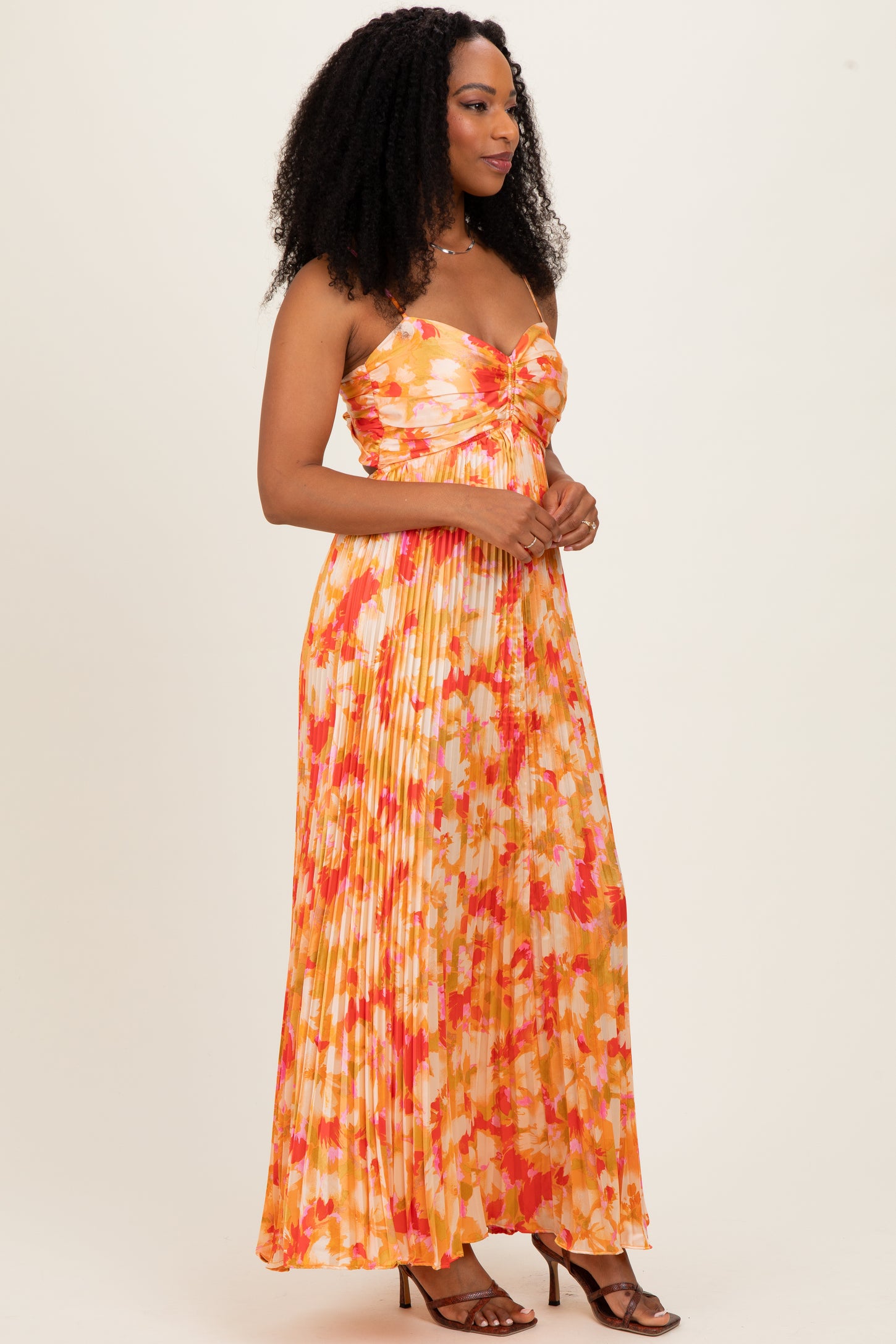 woman wearing an orange floral maxi dress with pleats and a twisted front detail, styled with strappy heels