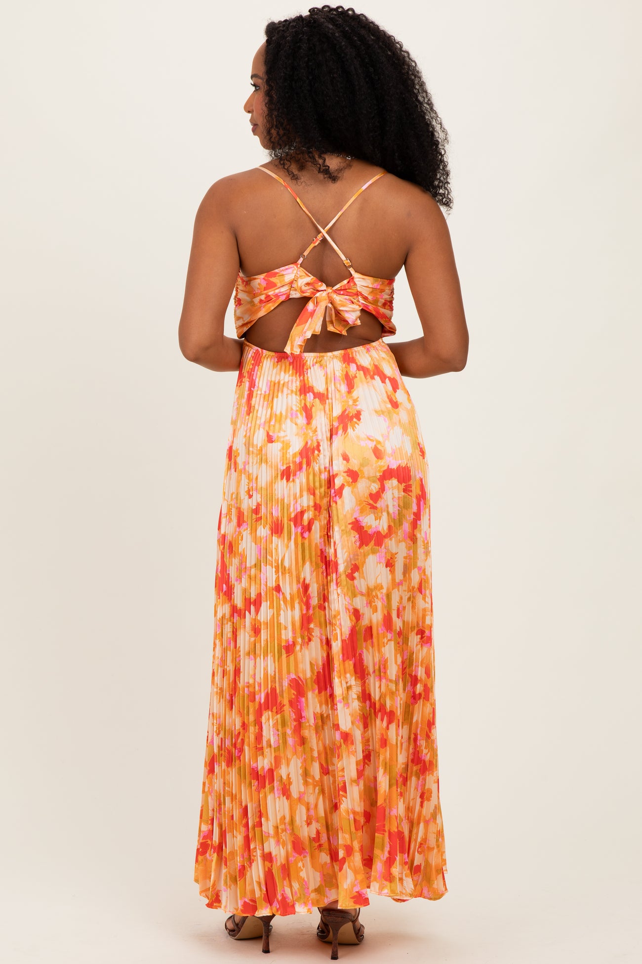 woman wearing a backless floral maxi dress with a tied detail and pleated skirt in shades of orange and pink