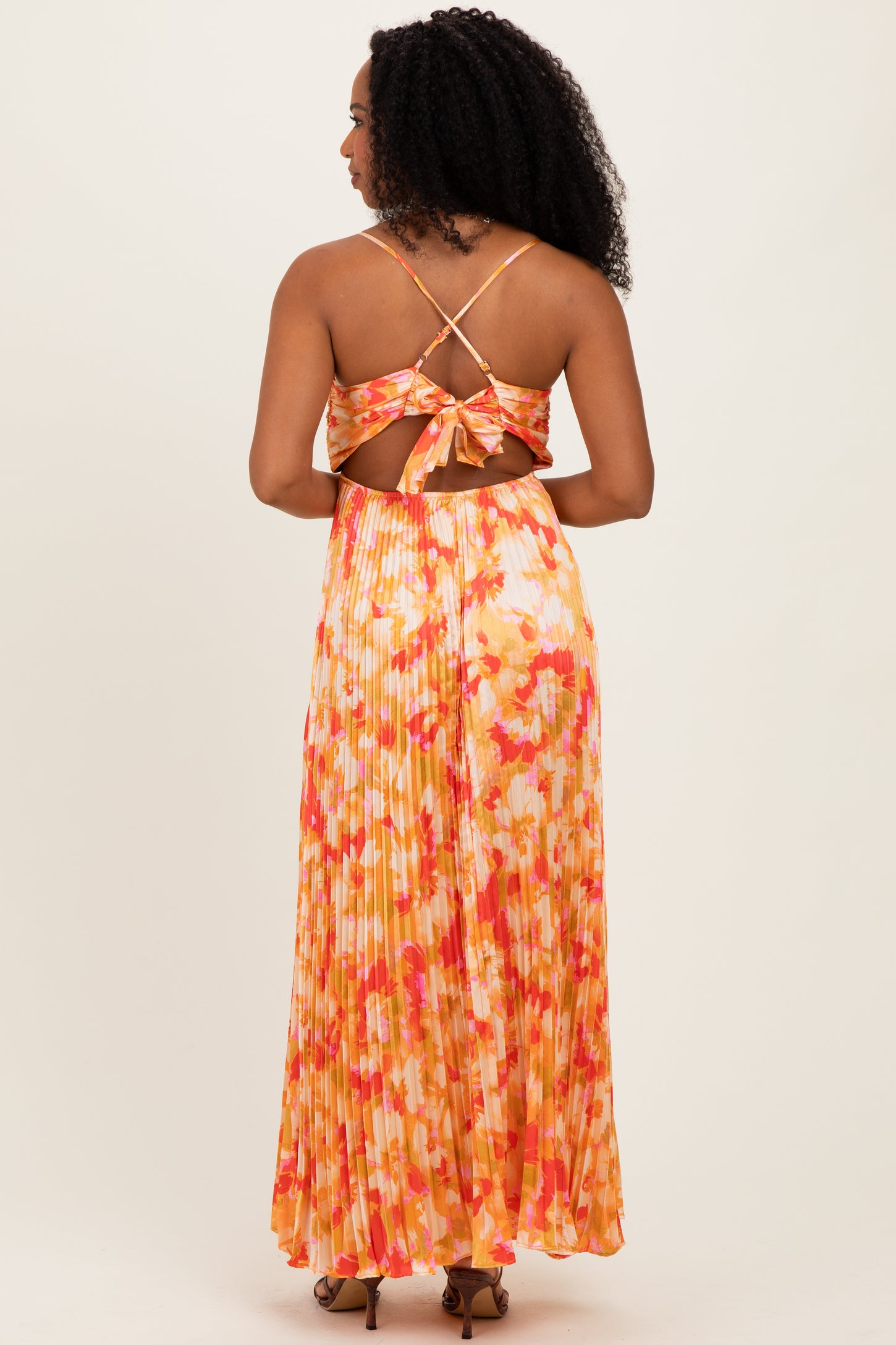 woman wearing a backless floral maxi dress with a tied detail and pleated skirt in shades of orange and pink