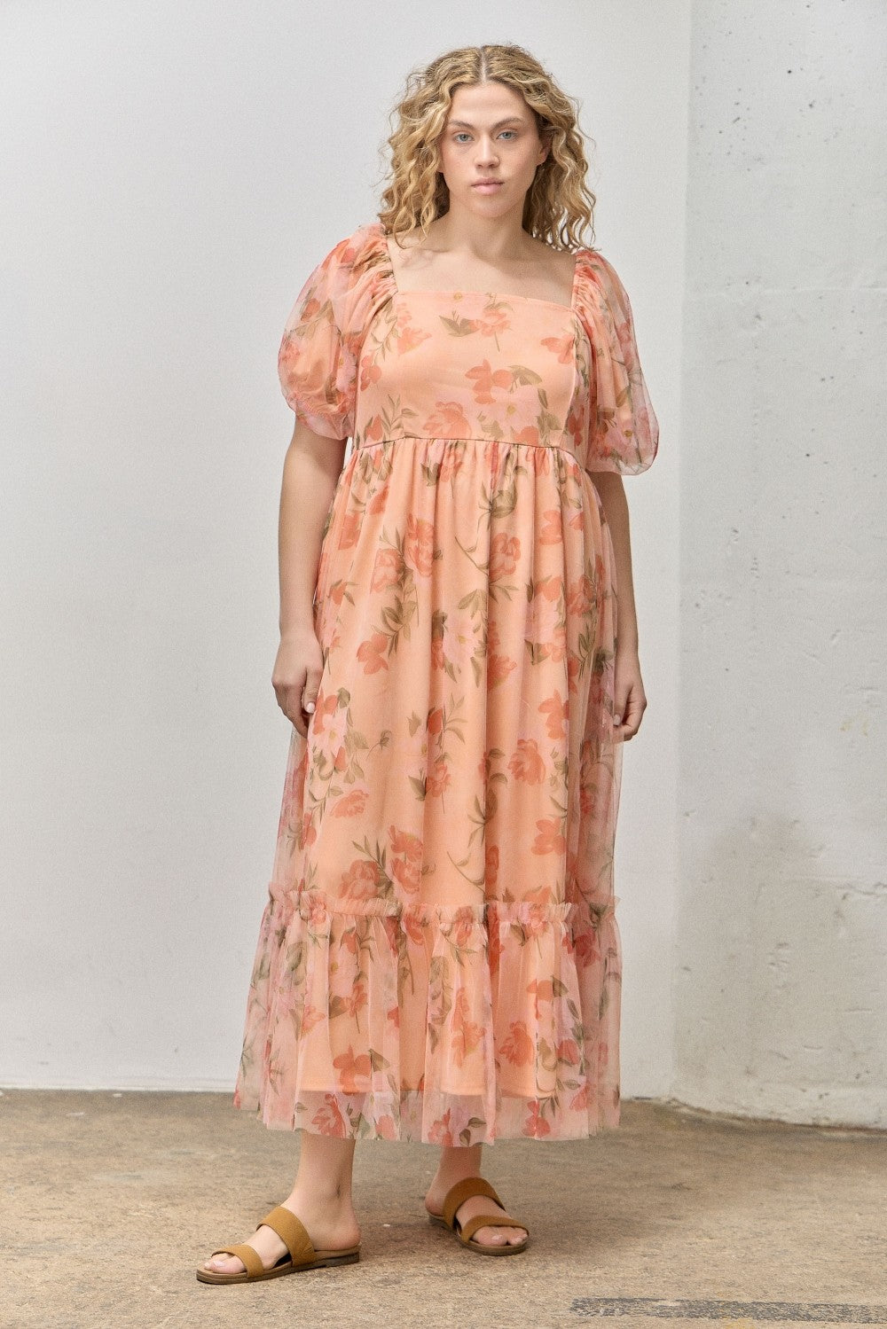 woman wearing a peach floral maxi dress with puff sleeves and a tiered hem, standing indoors on a concrete floor