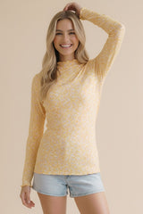 woman wearing a yellow long sleeve floral top and light denim shorts, smiling with one hand raised