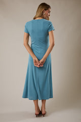 woman wearing a short sleeve blue midi dress, facing away with her hands behind her back