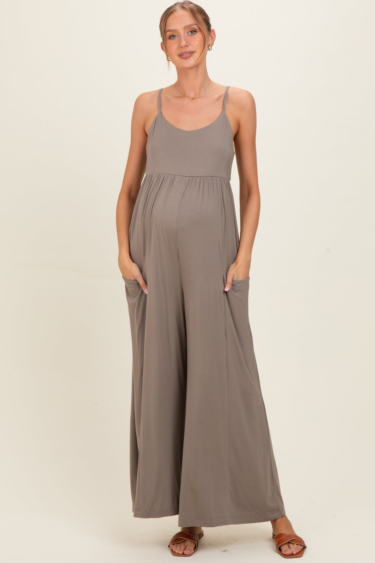 woman wearing a taupe maternity jumpsuit with pockets and adjustable straps, showcasing her baby bump