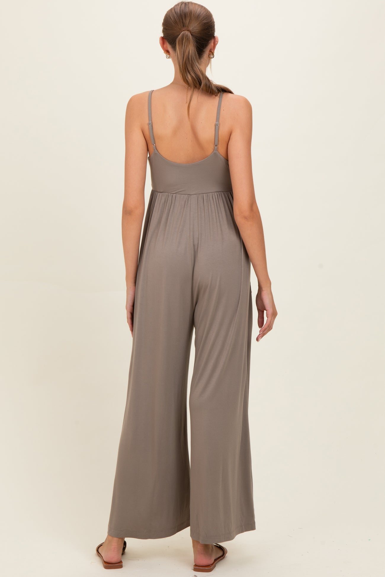 woman wearing a taupe maternity jumpsuit with a flowing wide leg and adjustable straps, showing the back view
