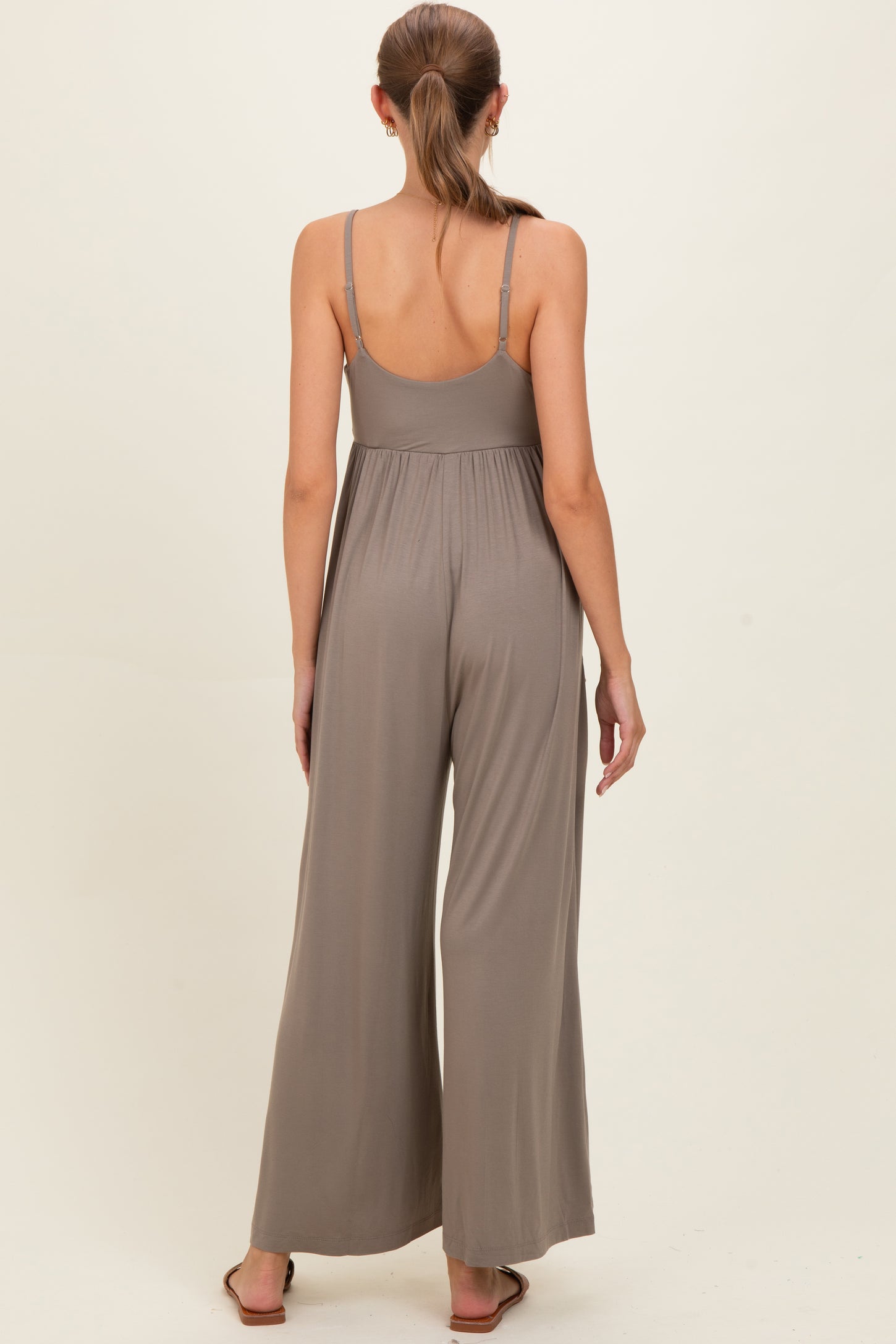 woman wearing a taupe maternity jumpsuit with a flowing wide leg and adjustable straps, showing the back view