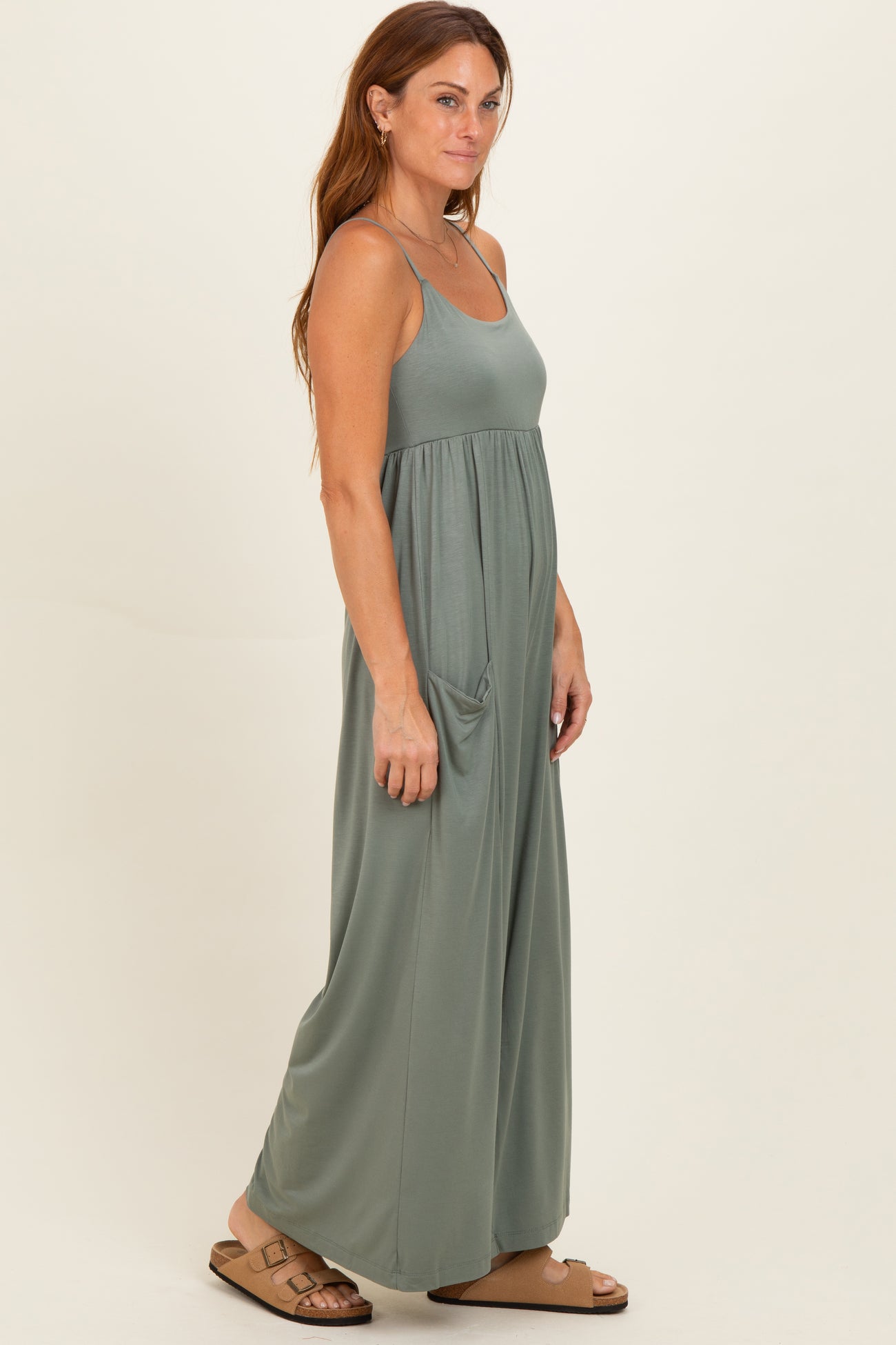 woman wearing a sage green maxi dress with thin straps and side pockets, standing in profile