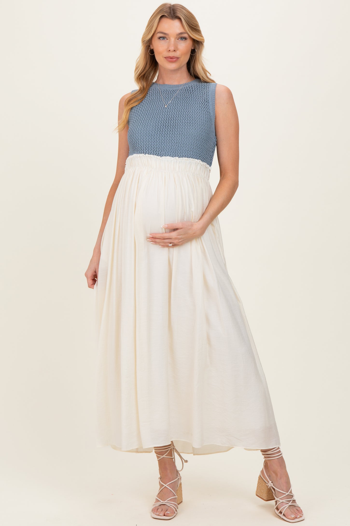 woman wearing a blue knit top and a flowing cream maternity dress, holding her baby bump