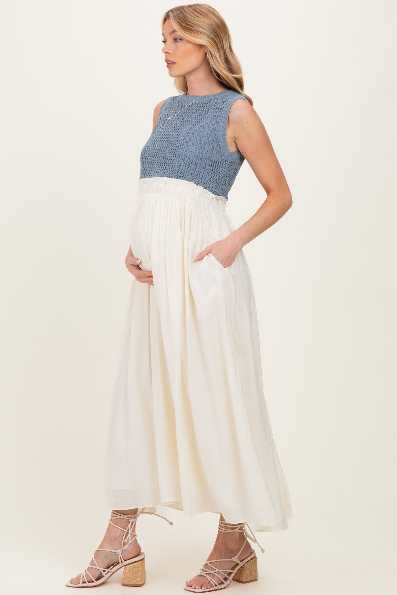 woman wearing a blue knit maternity top and a flowy white skirt, cradling her baby bump