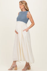 woman wearing a blue knit maternity top and a flowy white skirt, cradling her baby bump