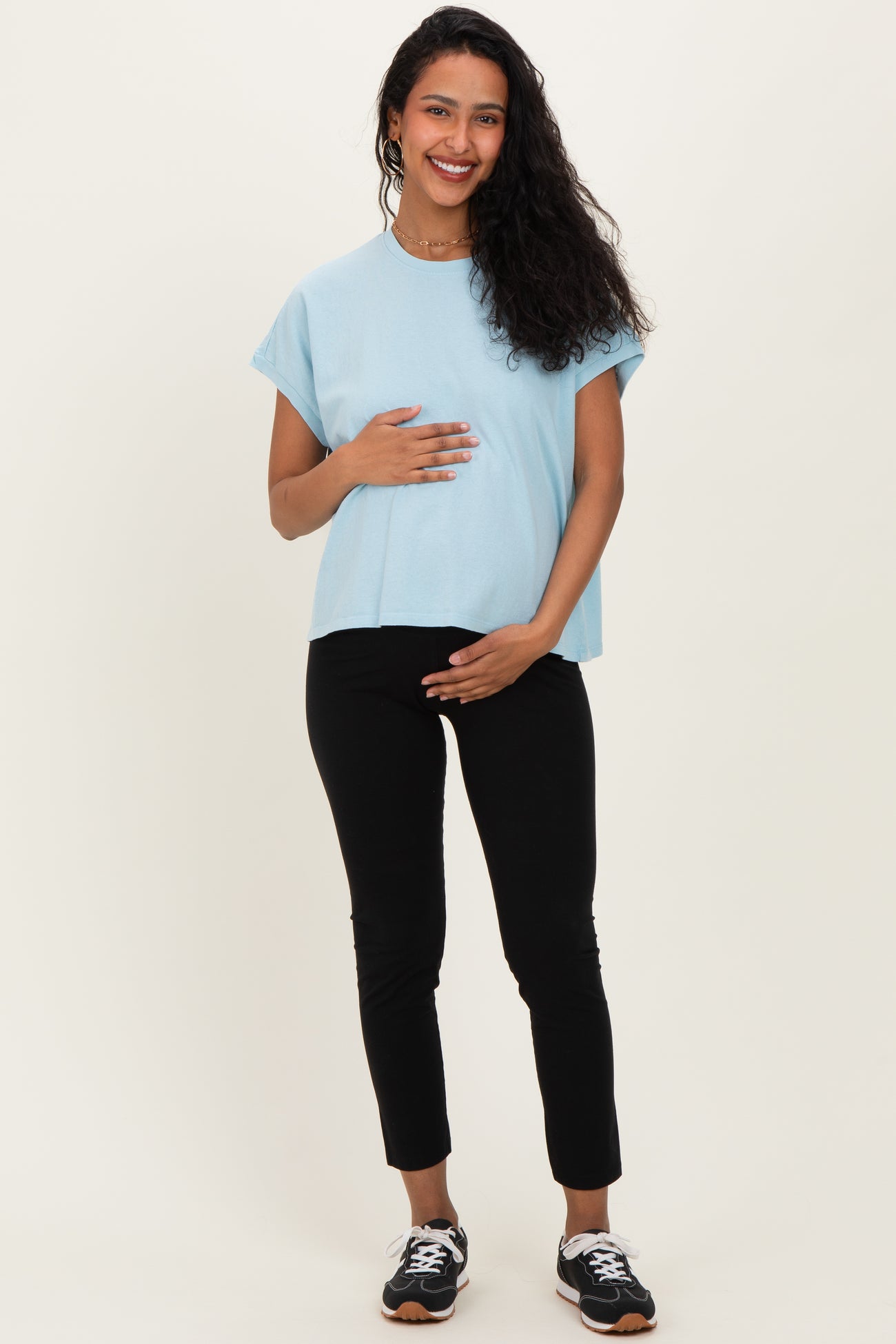 woman wearing a light blue maternity top and black maternity pants, holding her baby bump