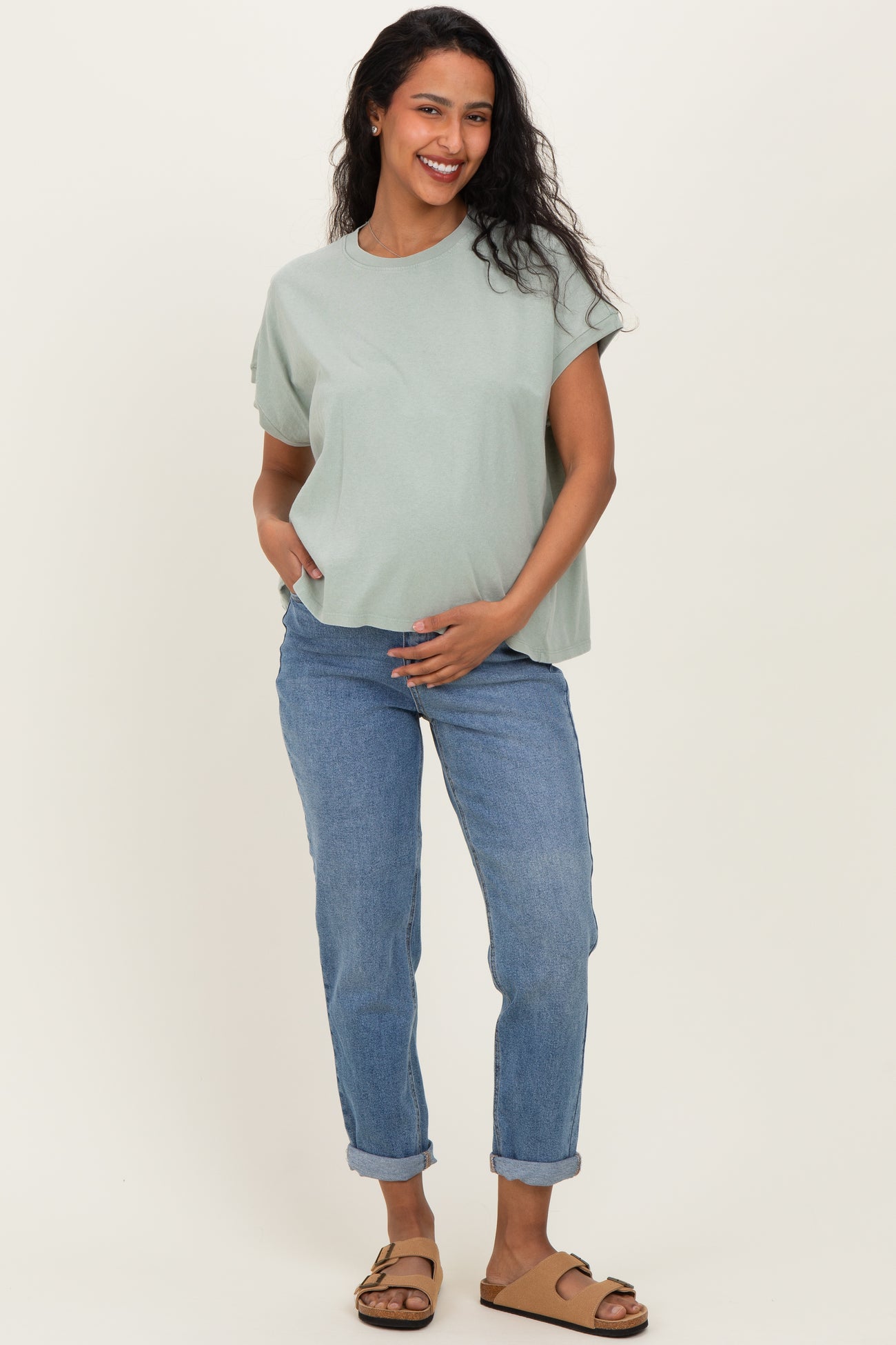 woman wearing a light green maternity top and blue rolled-up jeans, holding her baby bump