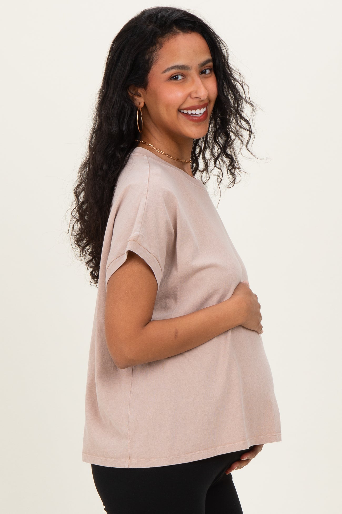 woman wearing a light pink maternity top and holding her baby bump, with a smile and curly hair