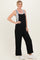 woman wearing a black sleeveless jumpsuit over a gray tank top, standing with one hand on her hip and wearing white sneakers