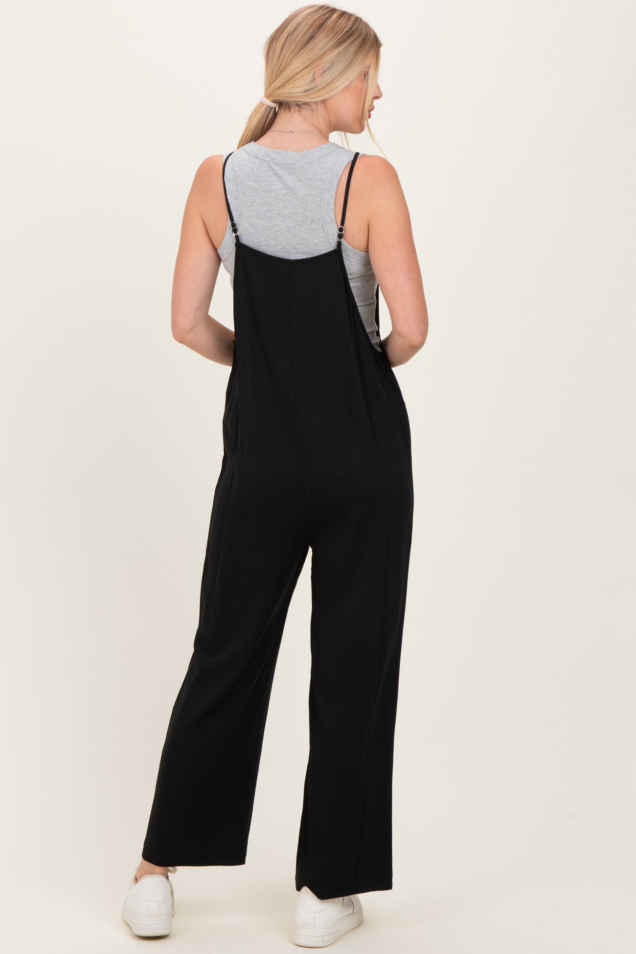 woman wearing a black jumpsuit over a gray tank top, viewed from the back, with straight leg fit and spaghetti straps