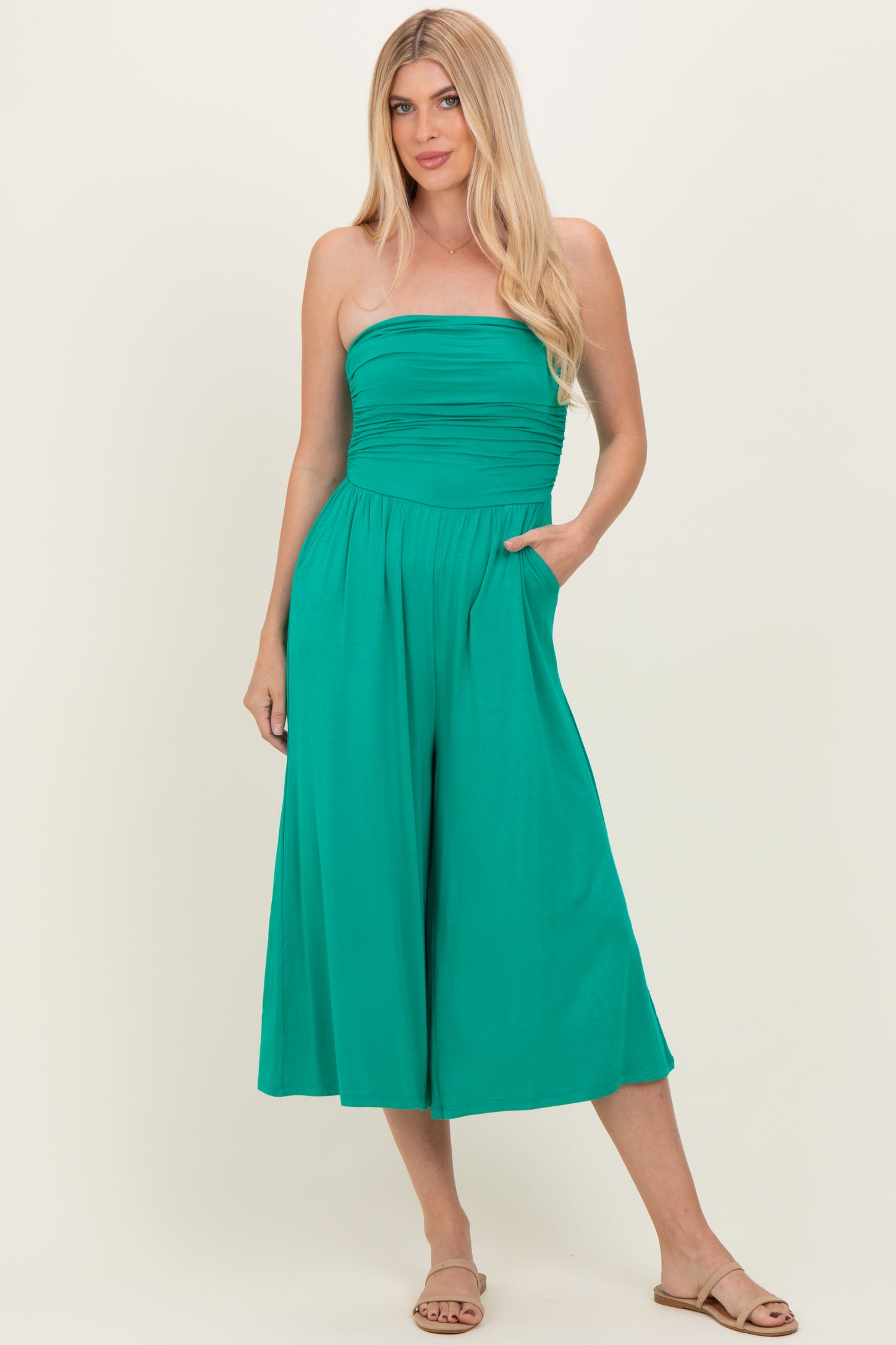 woman wearing a strapless teal jumpsuit with a ruched bodice and wide legs, posing with hands in pockets