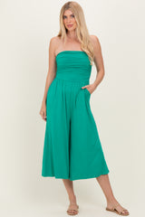 woman wearing a strapless teal jumpsuit with a ruched bodice and wide legs, posing with hands in pockets