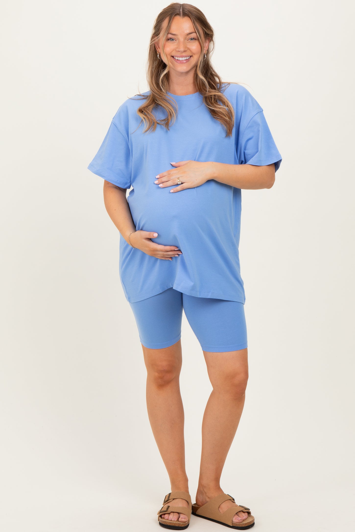 woman wearing a light blue oversized maternity top and matching bike shorts, gently cradling her baby bump