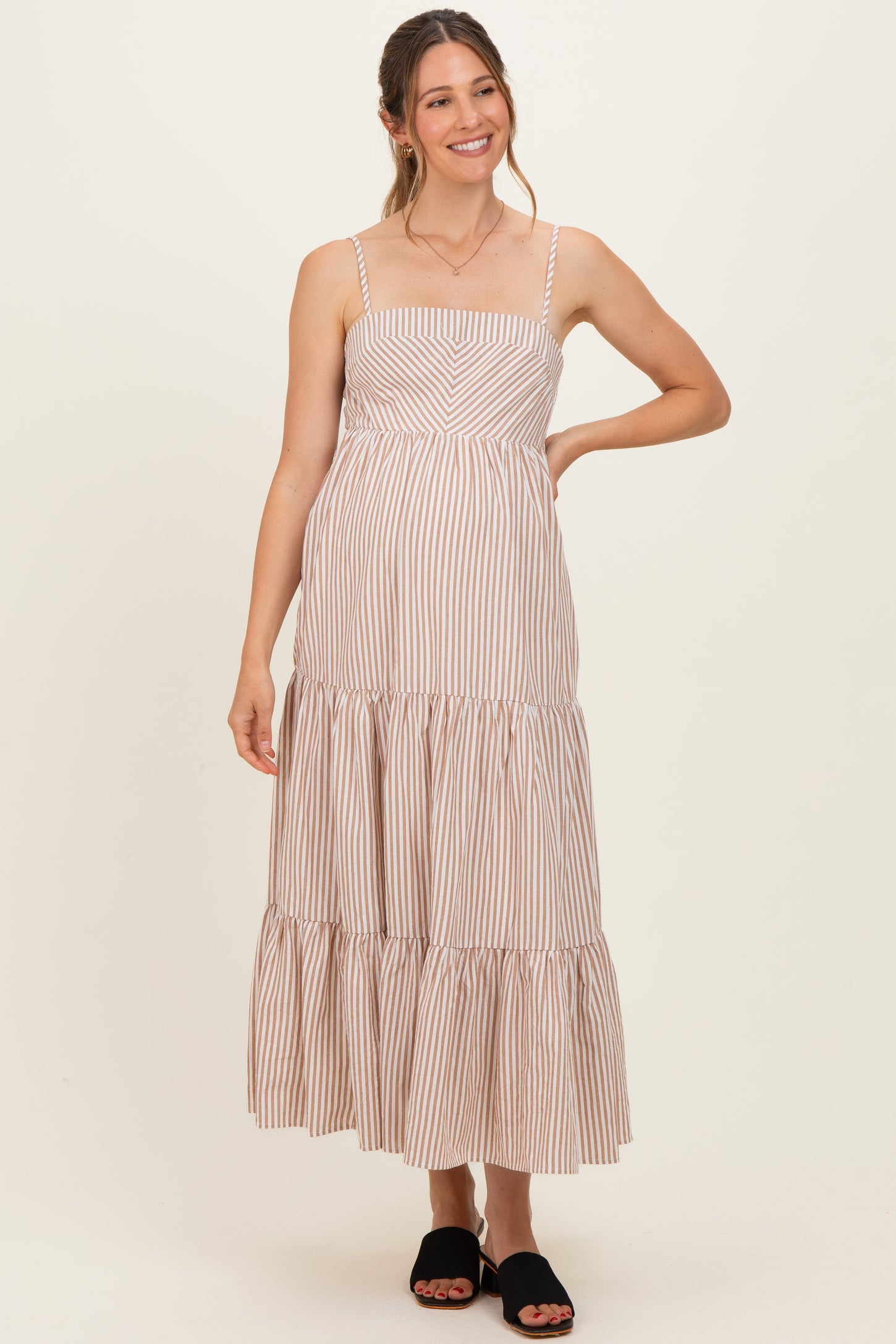 woman wearing a beige and white striped maternity maxi dress with spaghetti straps and tiered hem, smiling and standing in front of a plain background