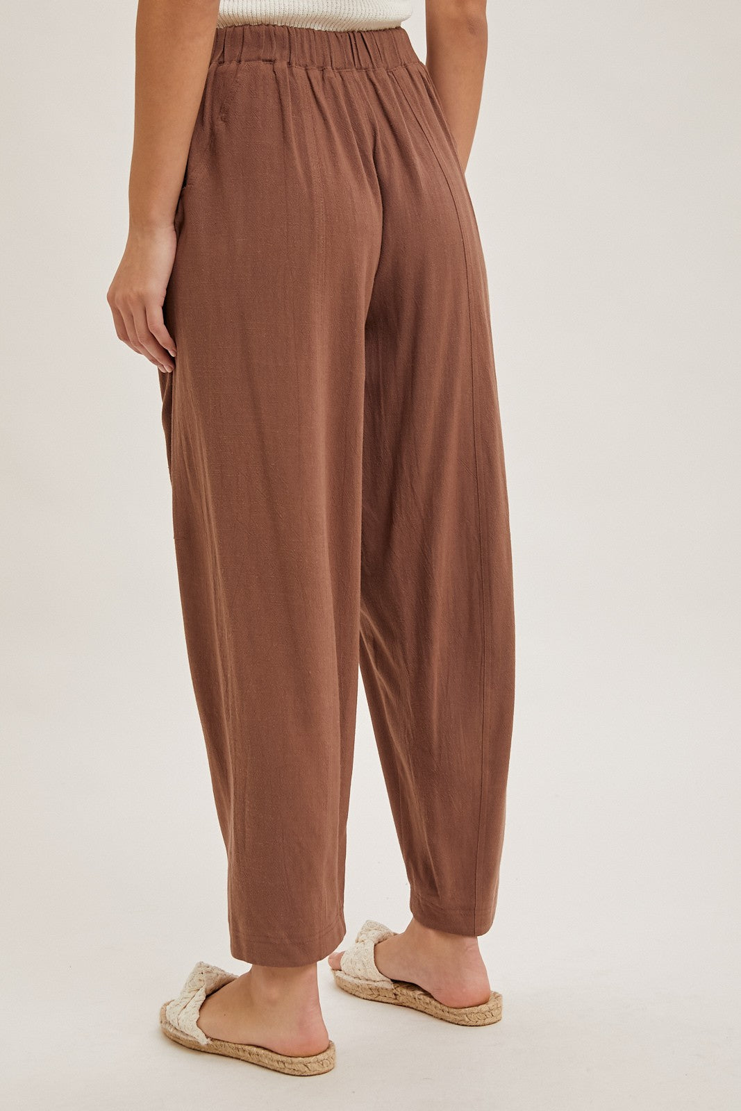 woman wearing brown wide-leg pants with an elastic waistband, standing barefoot with a light background