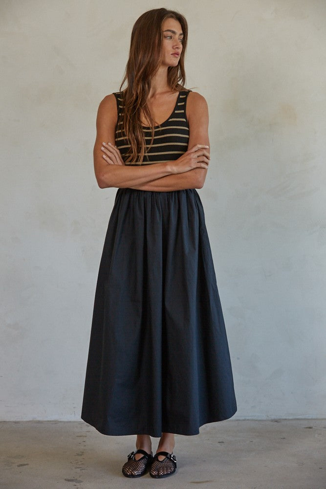 woman wearing a black and gold striped tank top and a long black skirt, standing with arms crossed