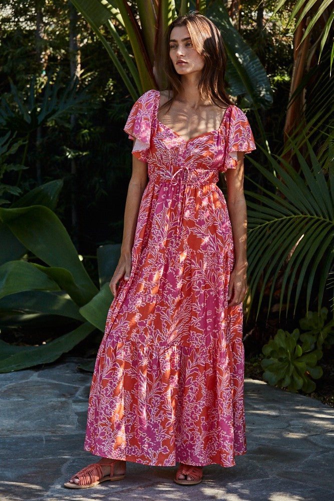 woman wearing a pink floral maxi dress with short ruffled sleeves standing outdoors among greenery