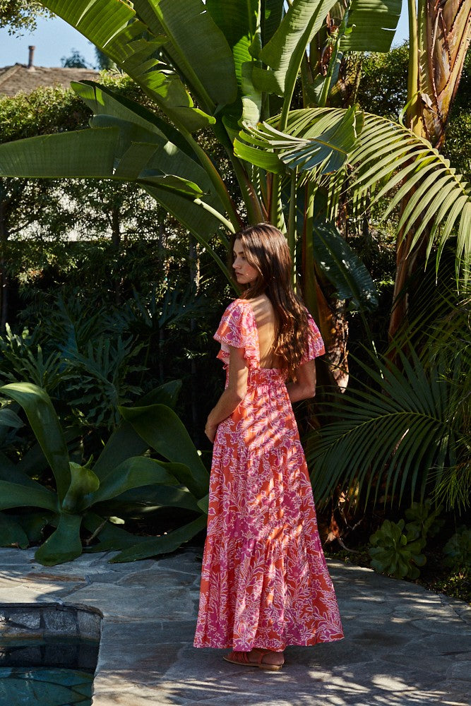 woman wearing a vibrant pink floral maxi dress with ruffled sleeves, standing in a tropical garden