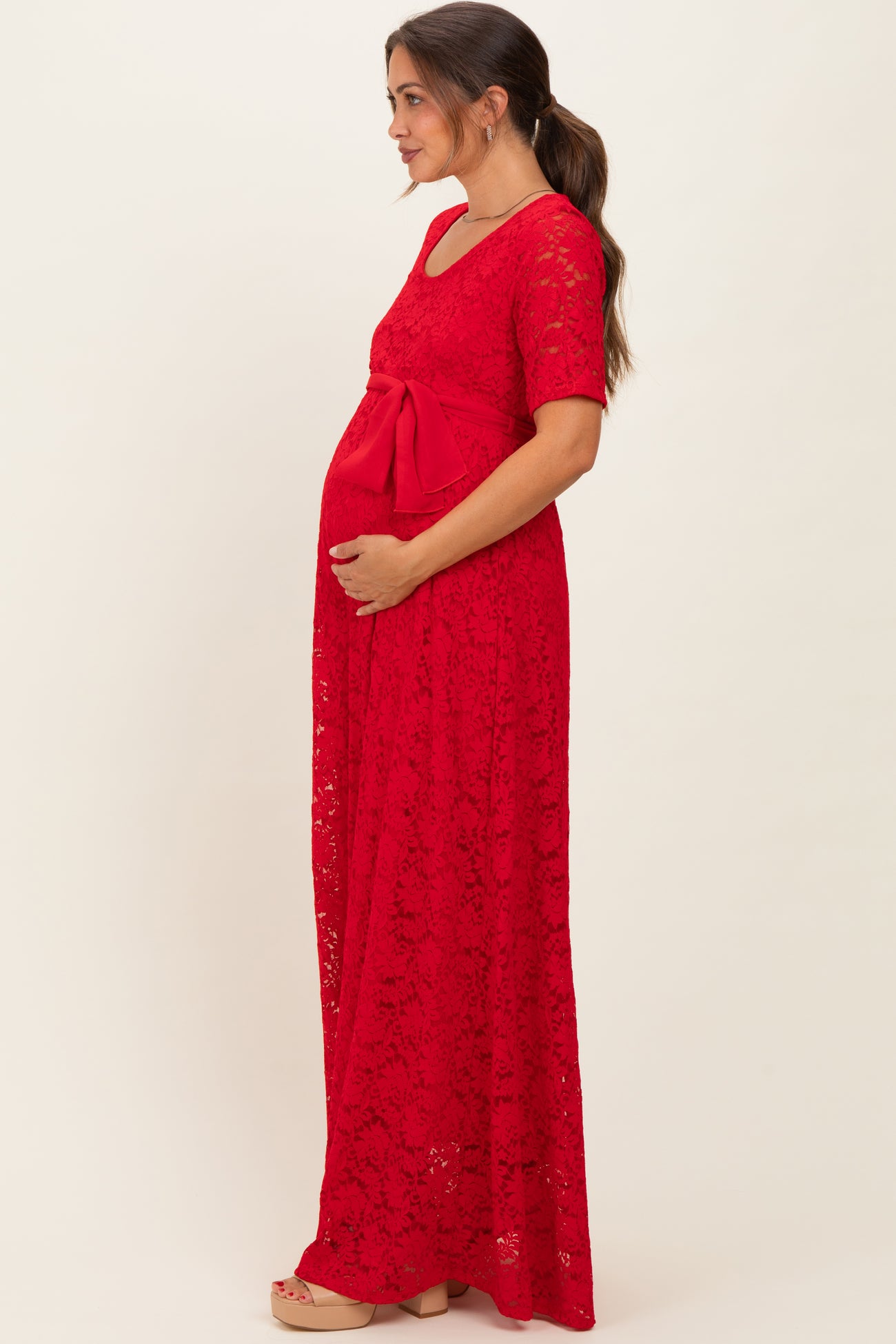 woman wearing a red lace maternity maxi dress with a bow detail and holding her baby bump