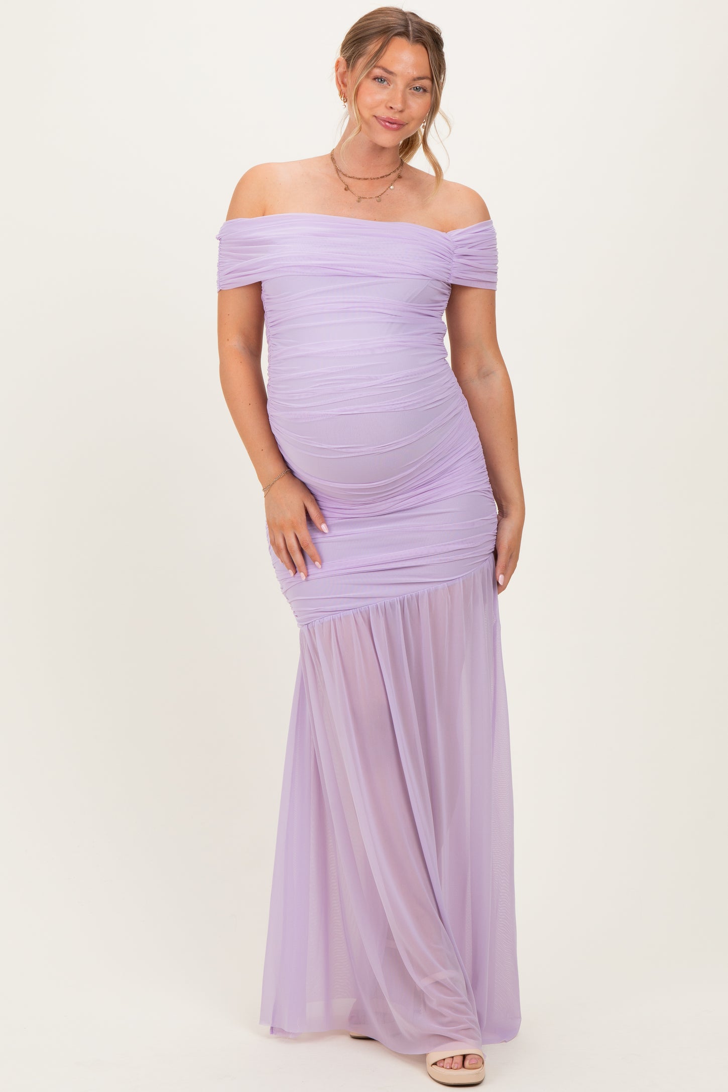woman wearing a lavender off-shoulder maternity gown with ruched details and a flowing skirt, showcasing her baby bump