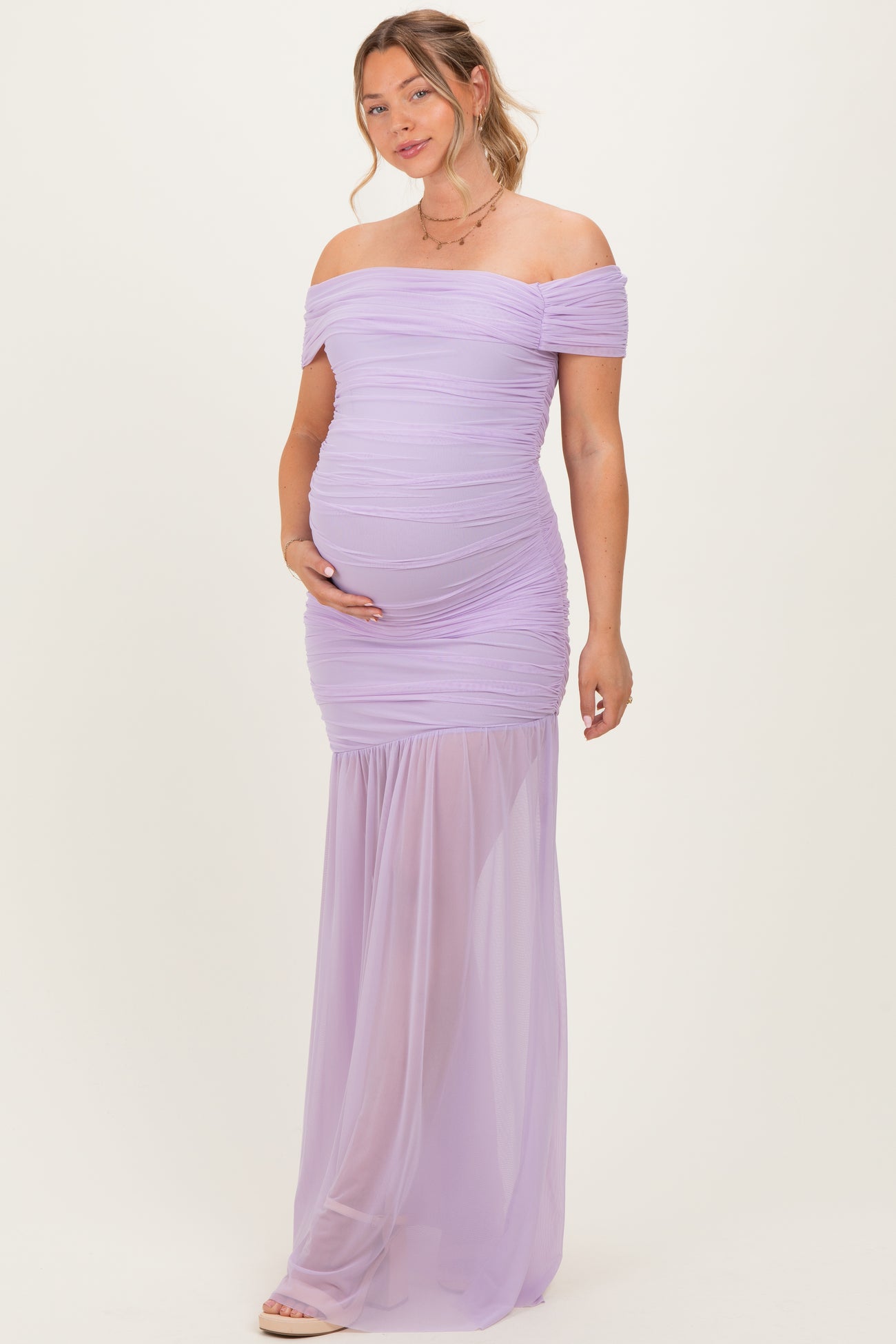 woman wearing a light purple off-the-shoulder maternity gown with ruching and a flowing skirt, holding her baby bump