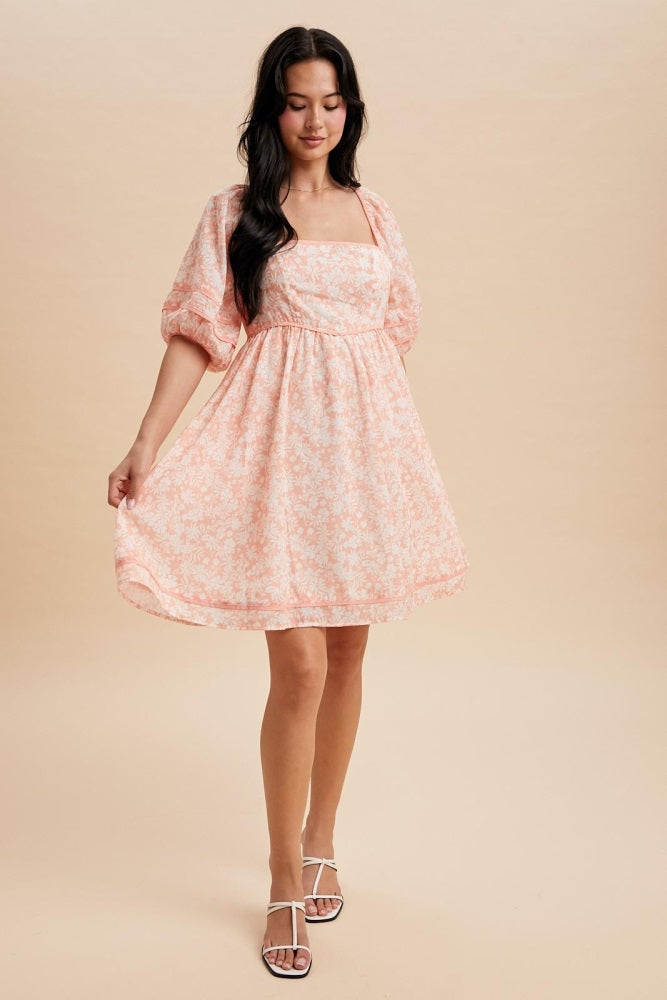 woman wearing a peach floral print short puff sleeve dress, holding the hem, standing against a neutral background