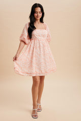 woman wearing a peach floral print short puff sleeve dress, holding the hem, standing against a neutral background