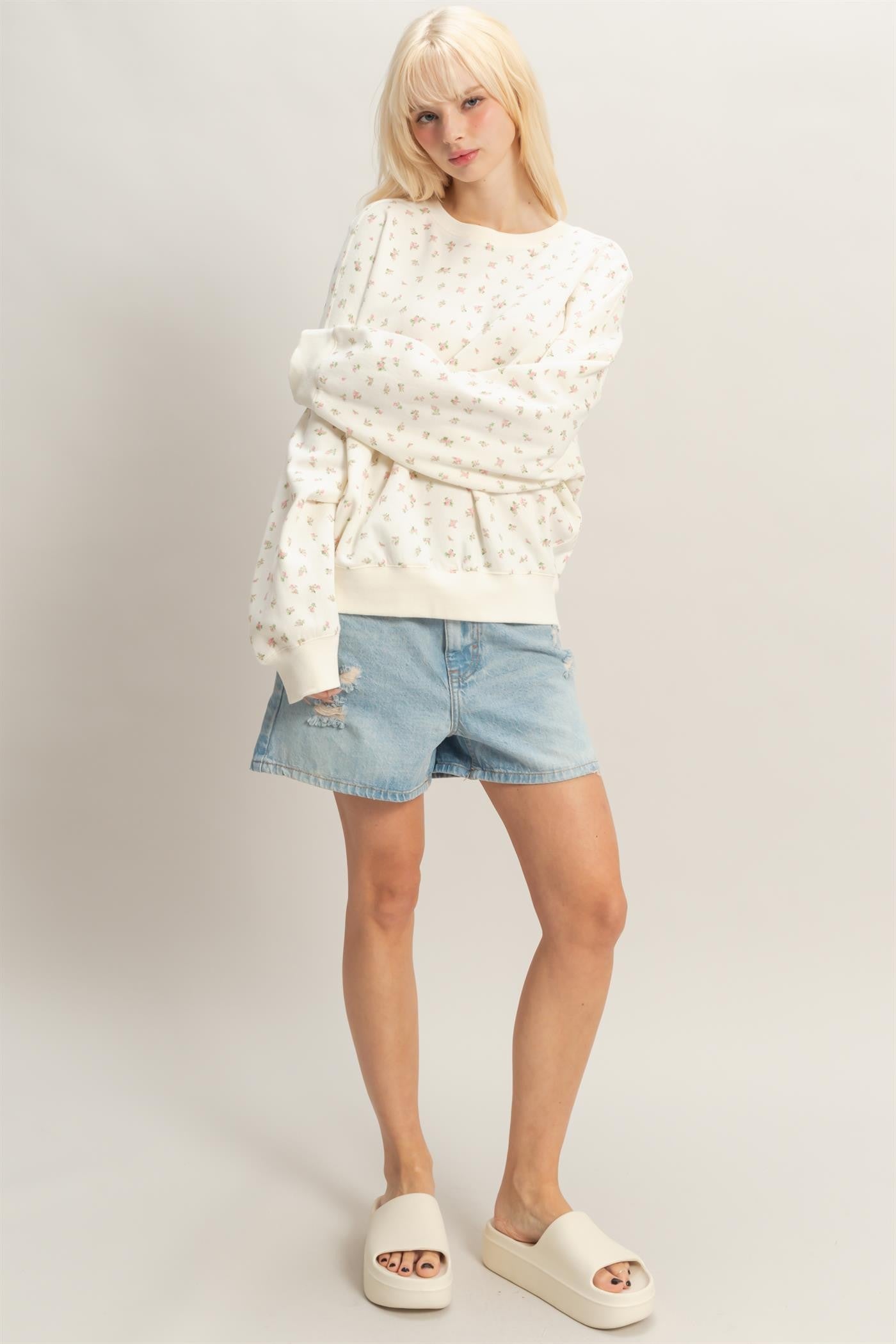 woman wearing a white floral sweatshirt and denim shorts, standing with a relaxed posture and wearing cream slide sandals