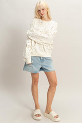 woman wearing a white floral sweatshirt and denim shorts, standing with a relaxed posture and wearing cream slide sandals