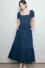 woman wearing a navy blue puff sleeve maxi dress with a square neckline and tiered detailing, standing with hands in pockets