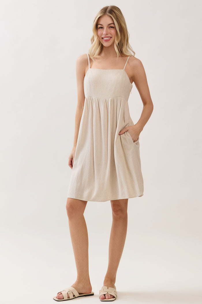 woman wearing a beige sleeveless sundress with a gathered waist and pockets, standing against a light background