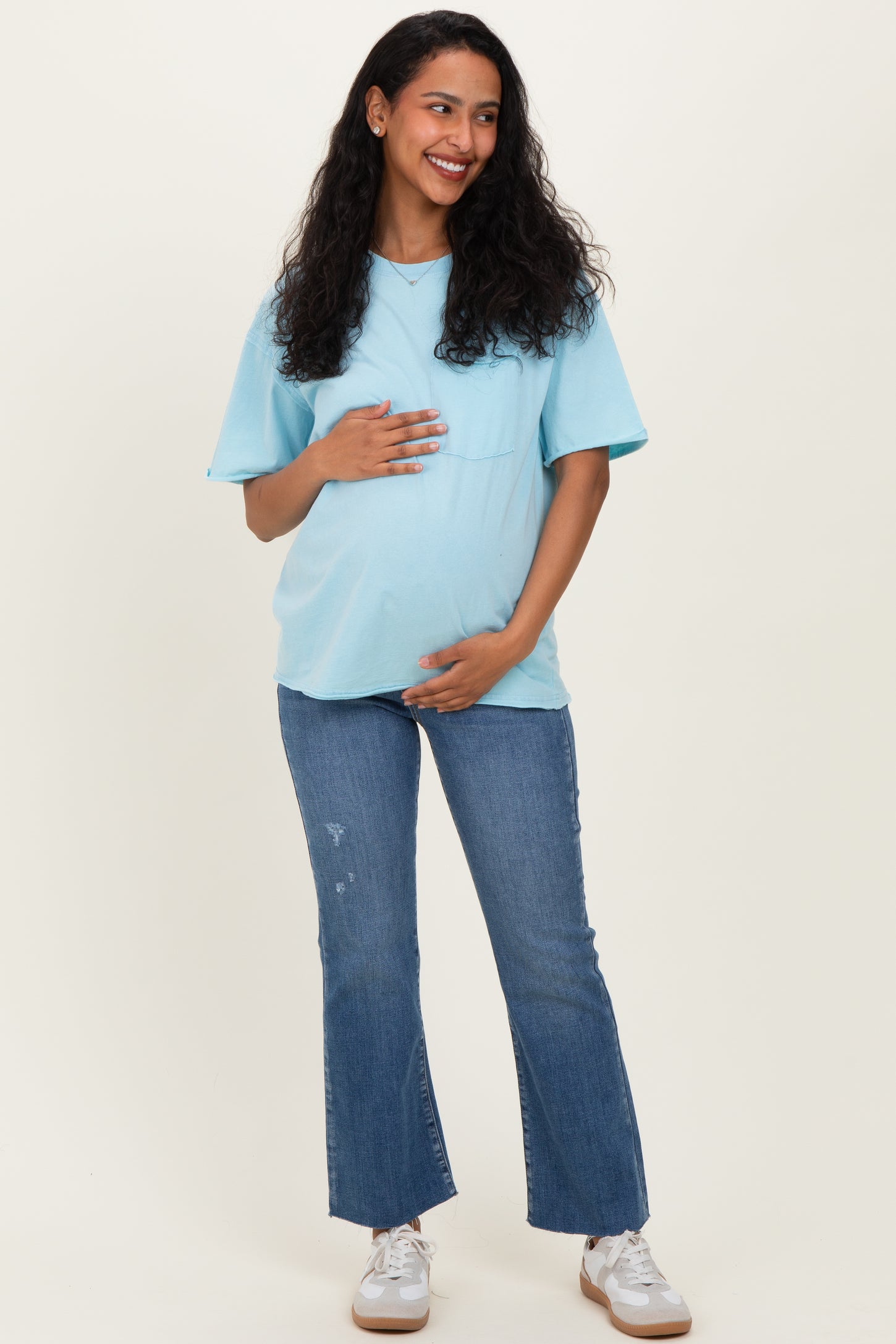 woman wearing a light blue short sleeve maternity top and distressed jeans, showing her baby bump with a smile