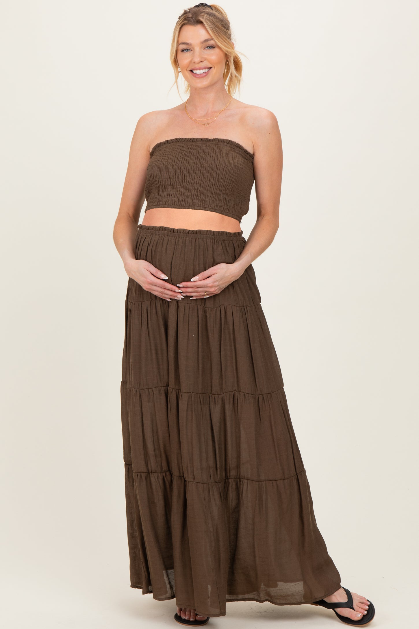 woman wearing a brown maternity two-piece outfit with a smocked strapless top and a flowy maxi skirt, holding her baby bump