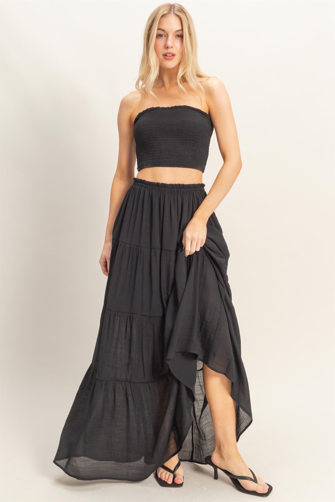 woman wearing a black smocked crop top and a black tiered maxi skirt with a high-low hemline and heeled sandals