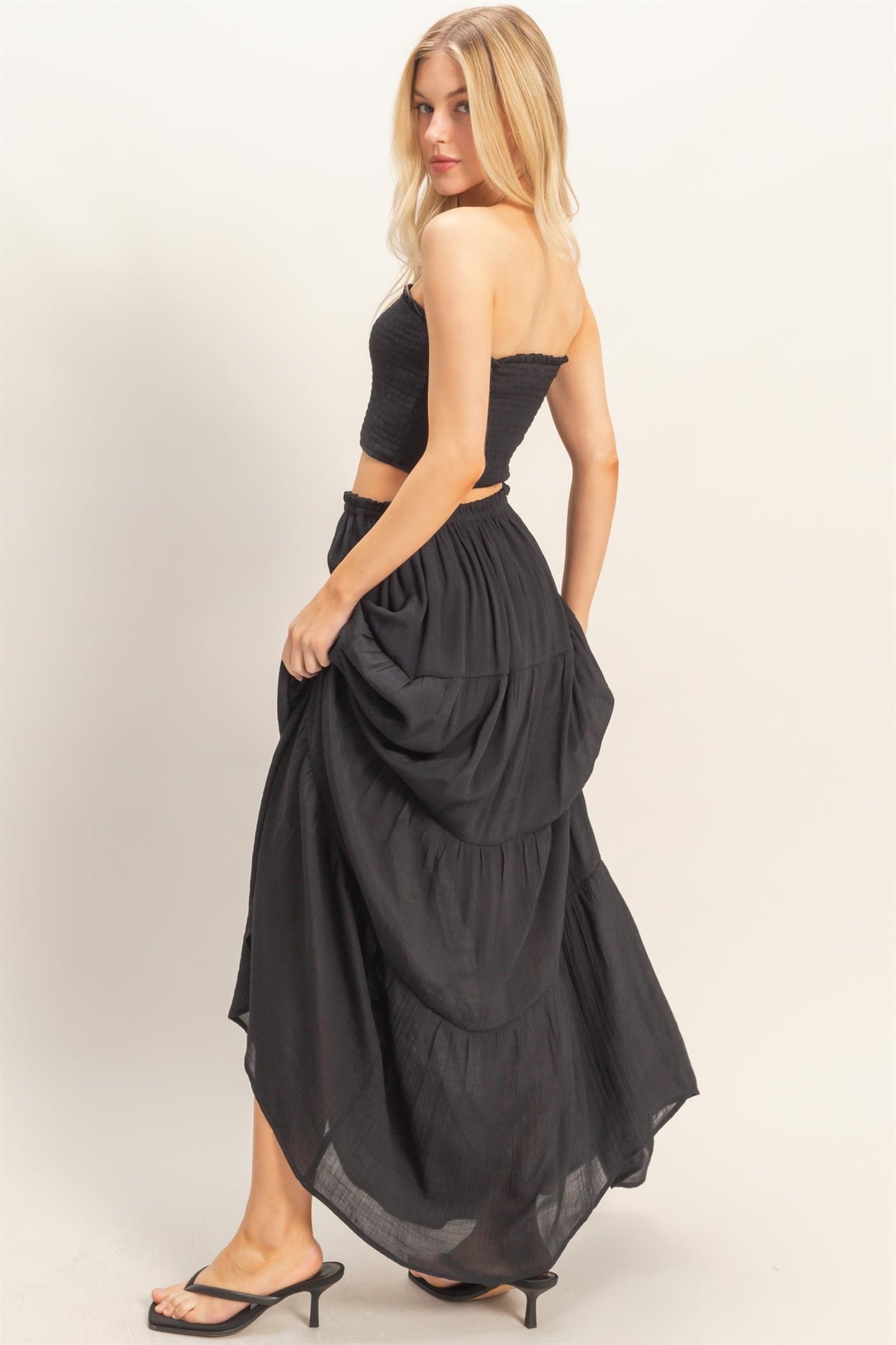 woman wearing a black strapless top and a flowy black maxi skirt with tiered details, standing with her back to the camera