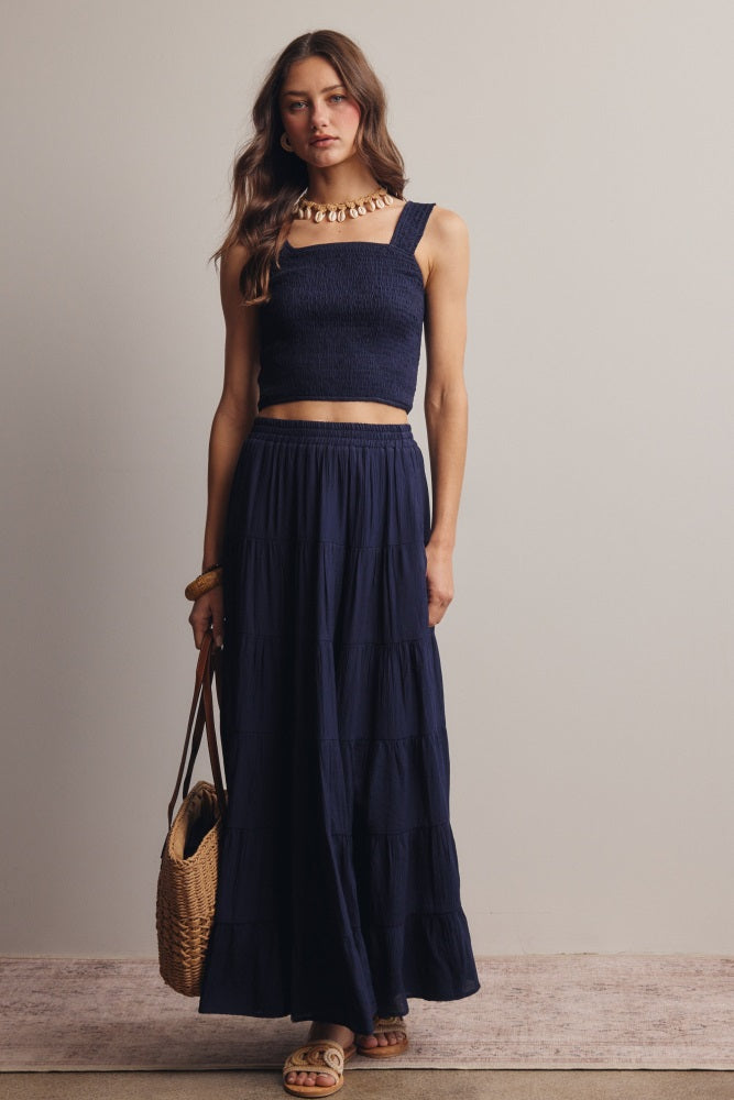 woman wearing a navy blue smocked crop top and a matching tiered maxi skirt, accessorized with a woven bag and jewelry