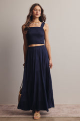 woman wearing a navy blue cropped smocked top and a flowy tiered maxi skirt, accessorized with a necklace and bag