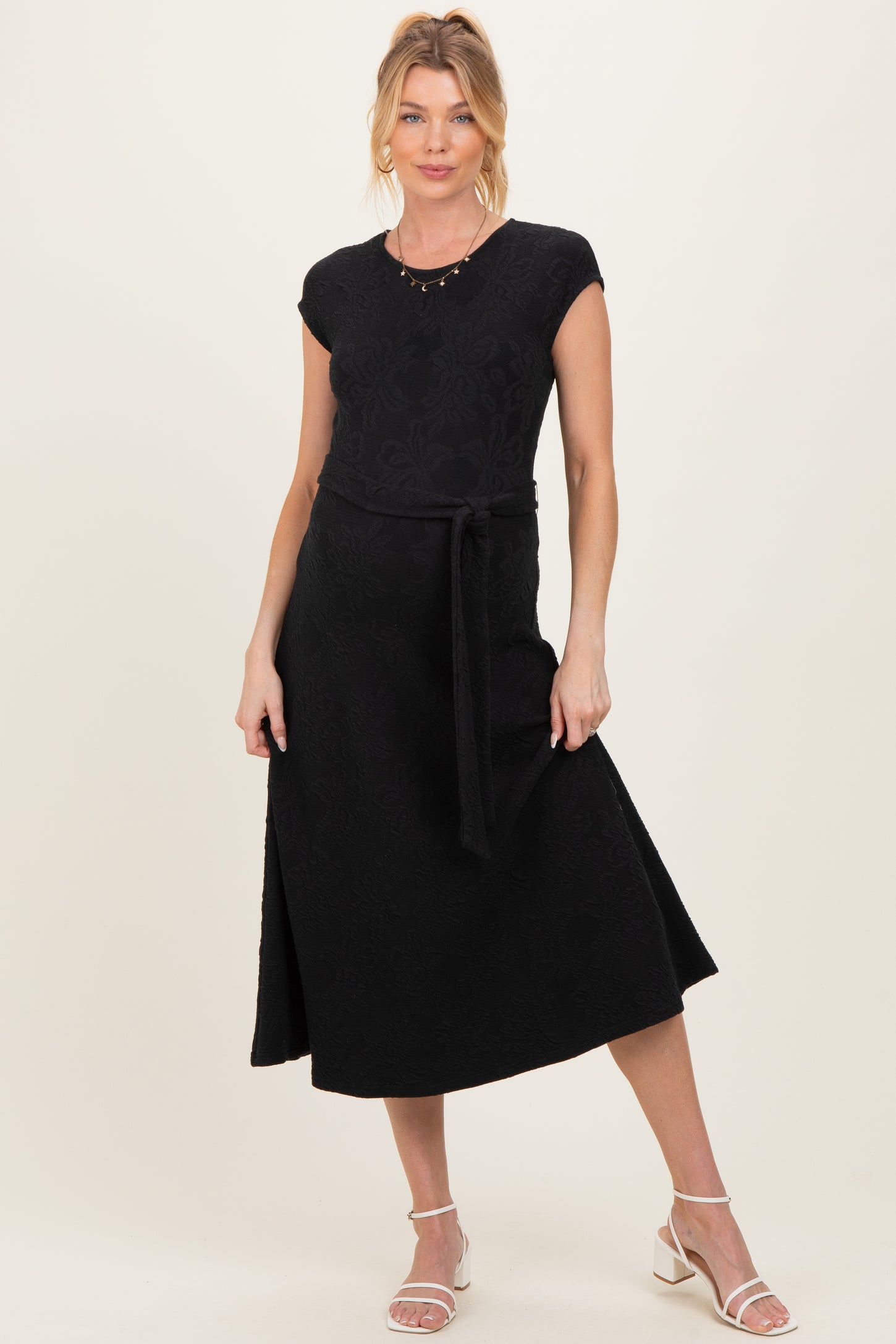 woman wearing a black maternity dress with a fitted top and a flowy skirt, accessorized with a belt and standing confidently