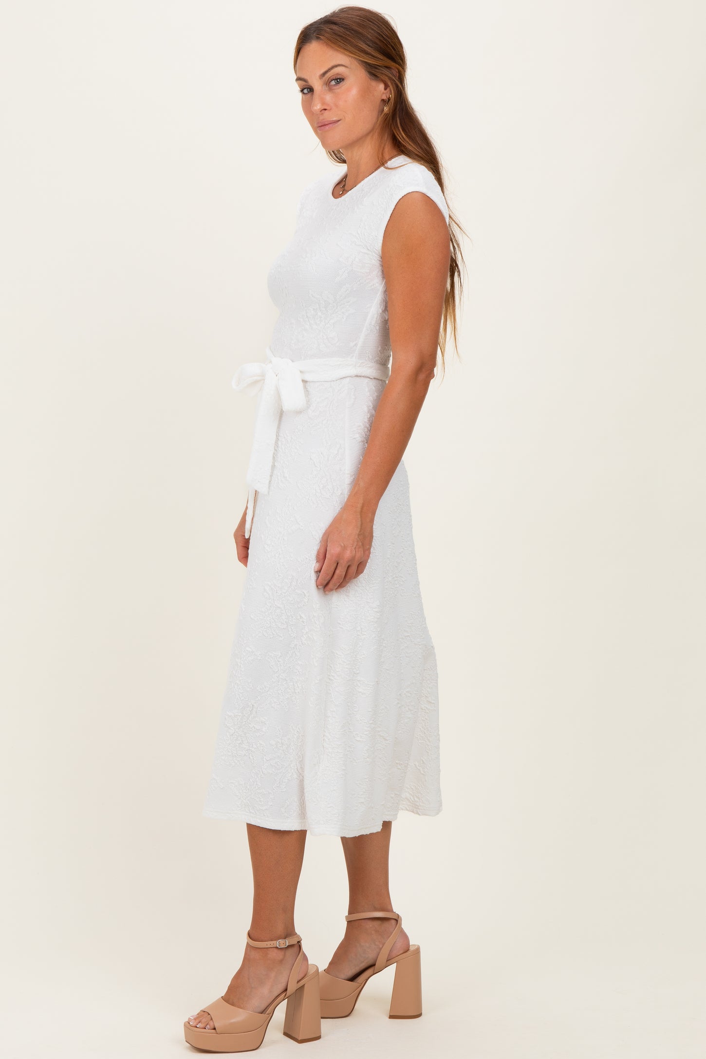 woman wearing a white sleeveless midi dress with a belt and a textured pattern, standing in a side profile pose