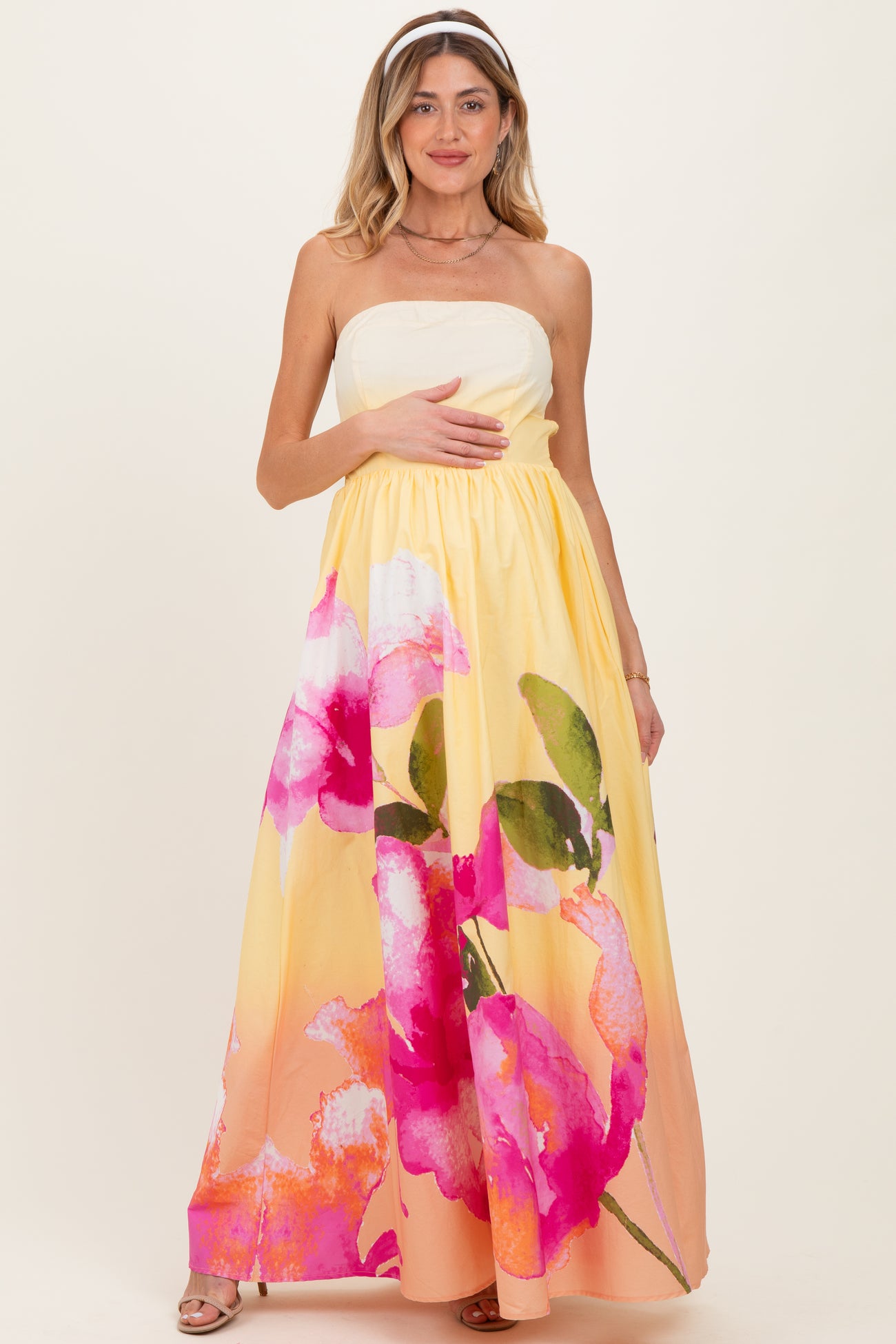 woman wearing a strapless maternity maxi dress with a yellow base and pink floral print, holding her baby bump