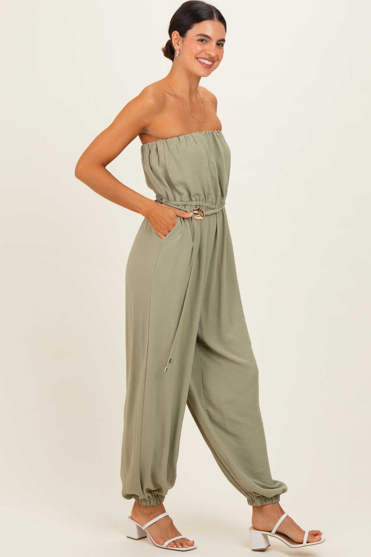 woman wearing a olive green strapless jumpsuit with a belted waist and elastic cuffs, posed sideways